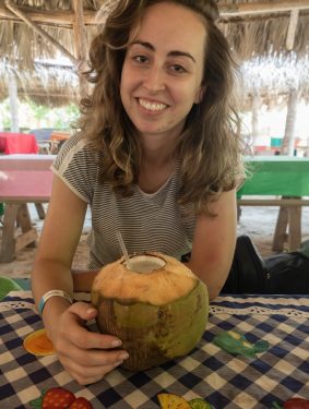 Roos de Leeuw with wavy brown hair and a black-and-white striped shirt holds a fresh green coconut with a white straw at an outdoor beachside table. She sits under a thatched-roof pavilion at a blue-and-white checkered tablecloth decorated with colorful fruit illustrations, with wooden tables and bright tropical daylight in the background.