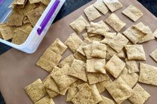 Batch of freshly baked keto walnut cracker thins, light brown squares on a mat and in a storage container.