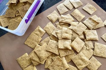 Batch of freshly baked keto walnut cracker thins, light brown squares on a mat and in a storage container.