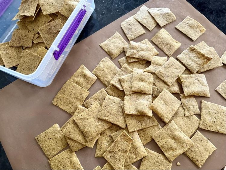 Batch of freshly baked keto walnut cracker thins, light brown squares on a mat and in a storage container.