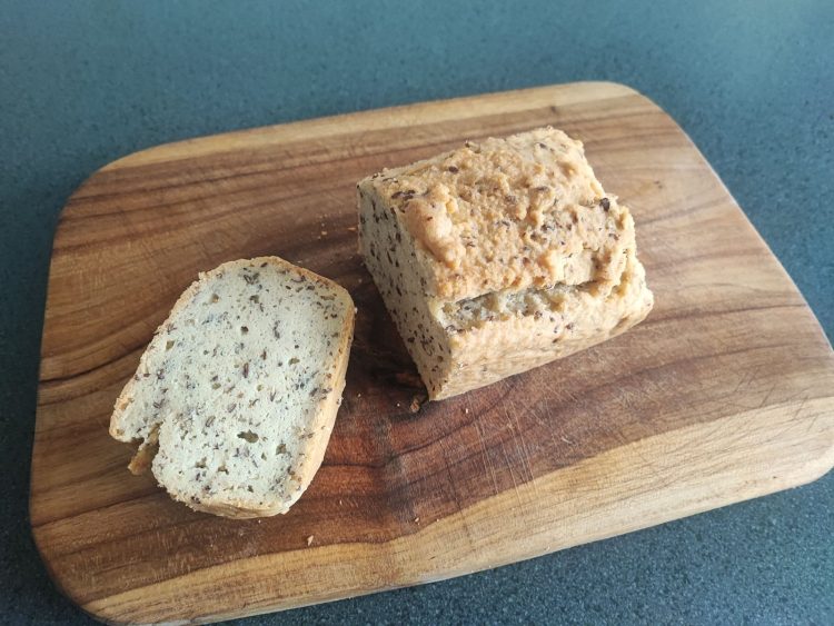A slice of seeded keto bread next to the loaf on a wooden board